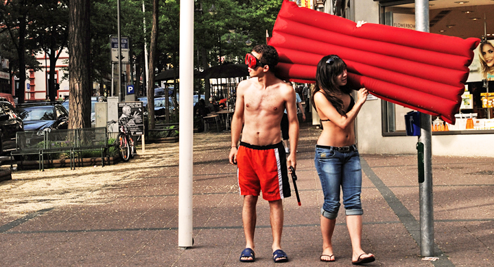 Ein junges Päärchen steht am Straßenrand, mit einer roten Luftmatratze in der Hand.